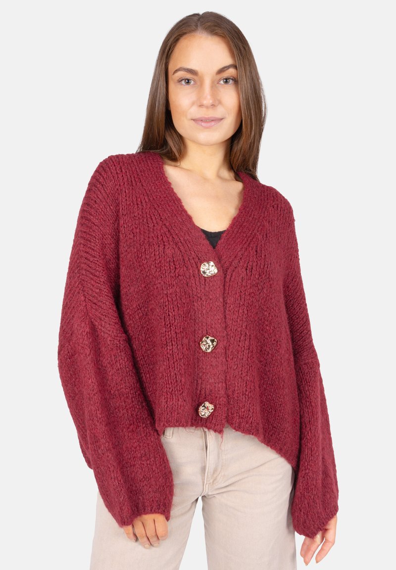 Femme portant un cardigan tricoté ample de couleur rouge profond avec trois boutons dorés décoratifs et un pantalon beige devant un fond uni.