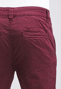 Pantalon en coton bordeaux avec une finition texturée, doté de deux poches arrière et de détails cousus le long de la ceinture et des coutures.