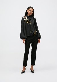 Černá halenka s dlouhým rukávem a květinovým vzorem, uvázaným výstřihem, spárovaná s černými slim-fit kalhotami a černými botami. Hladká textura látky.