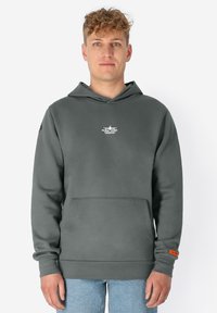 Jonge man met krullend haar draagt een donkergrijze hoodie met een voorzak en een klein wit vliegtuiglogo op de borst, gecombineerd met lichtblauwe jeans.