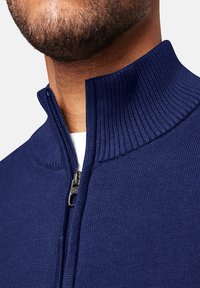 Pull en maille bleu marine avec un col montant et une fermeture à demi-zip. Présente une texture côtelée et une couleur unie, avec une doublure intérieure lisse.