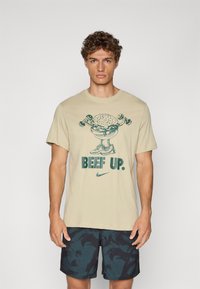 Beige bomulds T-shirt med et grønt grafik af en hamburger, der løfter vægte, med teksten "BEEF UP." Nike-logoet nederst.