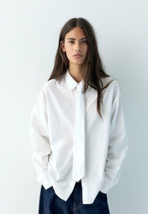 Młoda osoba o długich ciemnych włosach, ubrana w luźną białą koszulę i pasujący biały krawat, stojąca na tle jednolitego jasnego tła, z rękami w kieszeniach.