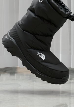 Schwarzer isolierter Winterstiefel mit dicker Sohle und The North Face-Logo, in der Luft über einem glatten Boden vor hellem Hintergrund gezeigt.