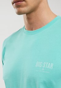 Tirkīza krāsas kokvilnas T-kreklu ar apaļu dekoltē, kas rotāts ar baltu logo tekstu "BIG STAR" un saukli. Gluda tekstūra, īsas piedurknes.