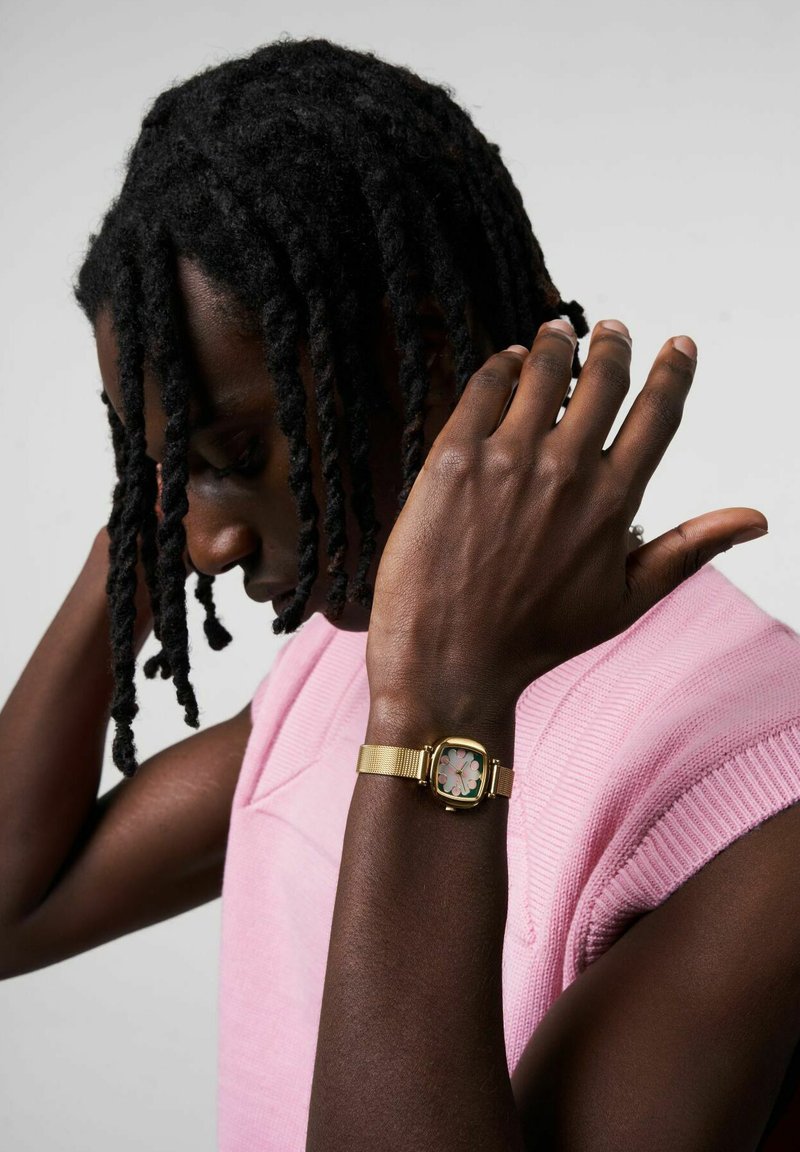 Montre avec un bracelet en maille dorée et un cadran carré, présentant un motif floral pastel. Le sujet porte un pull sans manches rose clair.