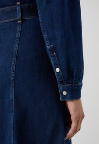 Mörkblå denimskjorta med långa ärmar, prydd med silverfärgade tryckknappar vid manschetterna och synlig sömnad längs sömmarna.