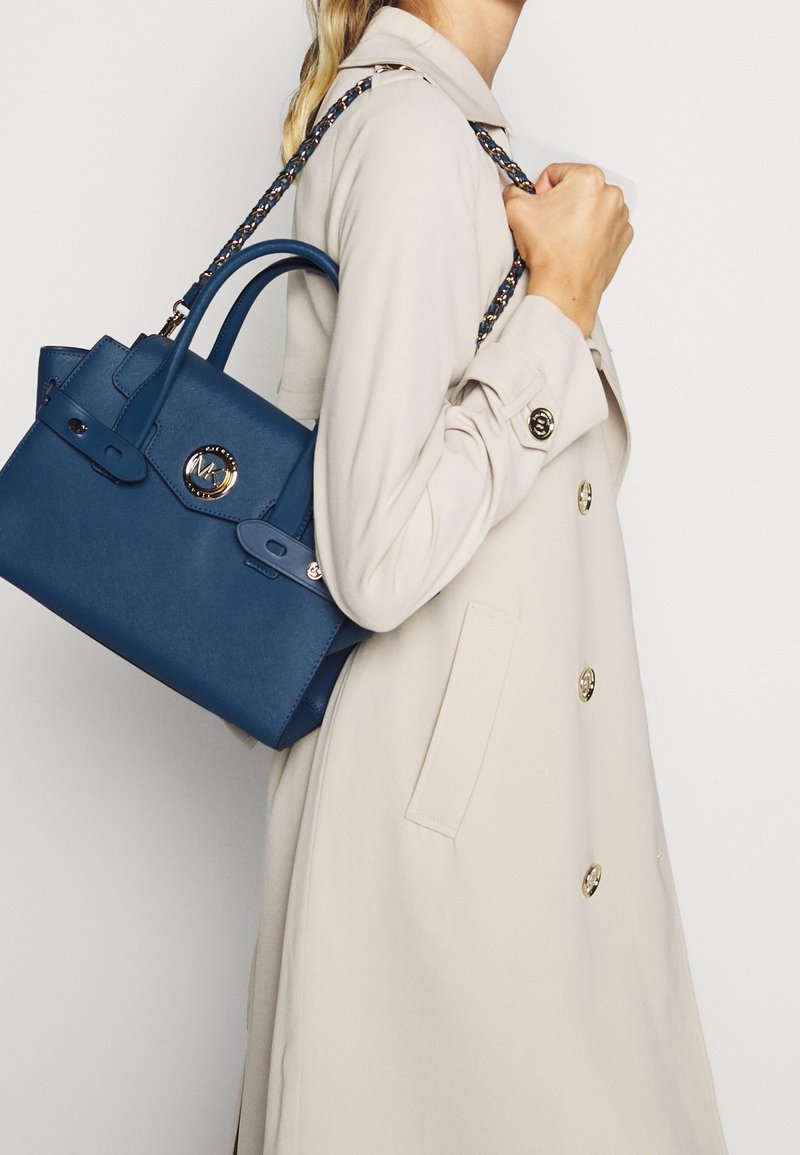 Femme en manteau beige portant un sac à main en cuir bleu avec un logo doré et une bandoulière tressée sur son épaule.
