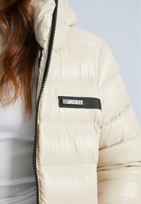 Puffjacke in hellbeige mit gestepptem Design, schwarzem Reißverschluss und einem schwarzen Etikett mit dem Text "JACKIT" auf der Vorderseite.
