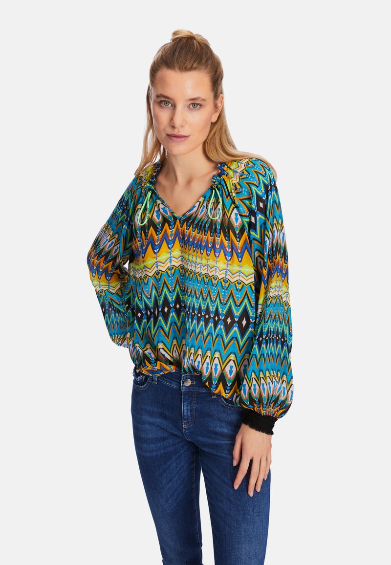 Colorful, patterned blouse featuring chevron stripes in blue, yellow, and black. Puff sleeves with elastic cuffs. V-neck design.
