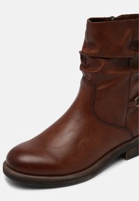 Bottine en cuir marron avec un design froncé, bout arrondi, fermeture éclair latérale et petit talon en bois. Surface texturée avec détails cousus.