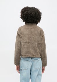 Osoba s kučeravými vlasmi, oblečená v hnedej flísovej bunde a svetlomodrých džínsach, otočená chrbtom na jednoliatom bielom pozadí.