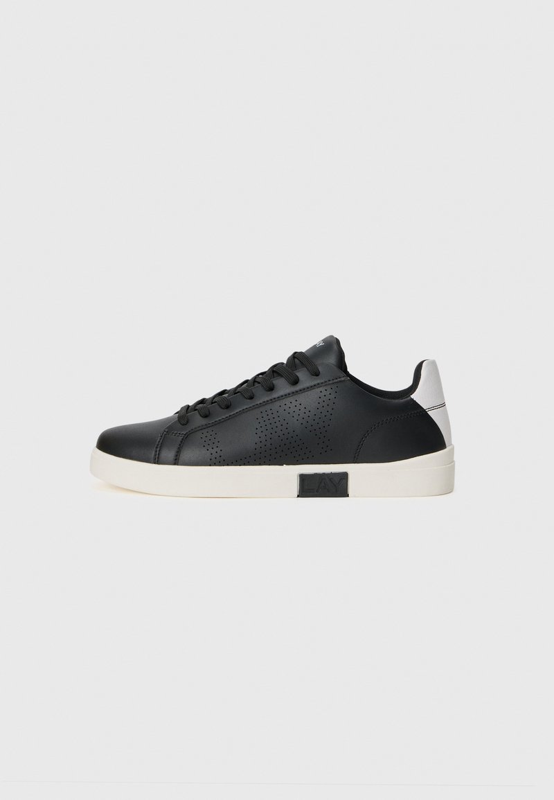 Zapatilla de piel negra con detalles perforados en los laterales, suela blanca y un logo sutil en el talón, exhibida sobre un fondo blanco liso.