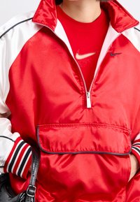 Person som bär en röd och vit Nike-jacka över en röd Nike-t-shirt, håller en svart texturerad handväska och har händerna i jackfickorna.