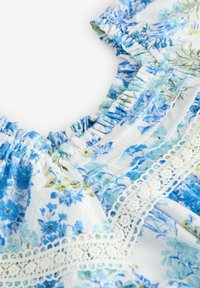 Lätt blommig blus i vitt med blå och gula blommönster. Har rynkad halsringning och intrikata spetsdetaljer.