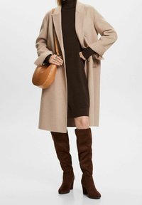 Cappotto di lana beige sopra un abito di maglia marrone scuro, abbinato a stivali al ginocchio in suede marrone e a una borsa a tracolla in pelle color beige. Design semplice ed elegante.