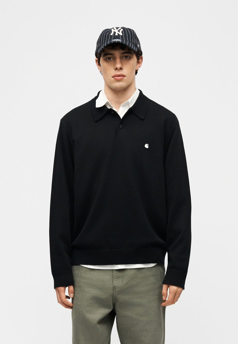 Junger Mann, der ein schwarzes Poloshirt mit langen Ärmeln, ein weißes Hemd darunter, olivgrüne Hose und eine marineblaue Baseballkappe mit Nadelstreifen trägt.