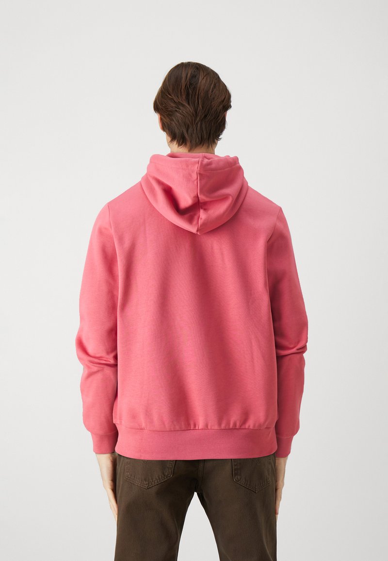 Hoodie en polaire rose avec capuche à cordon, poignets et ourlet côtelés. Texture lisse, coupe décontractée, associé à un pantalon brun. Vue de dos.