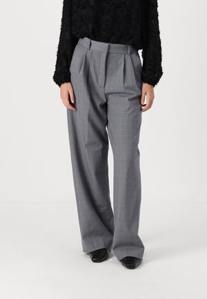 Femme portant un pantalon large à taille haute à carreaux gris avec des plis, associé à un haut texturé noir à manches longues et des chaussures noires à bouts pointus.