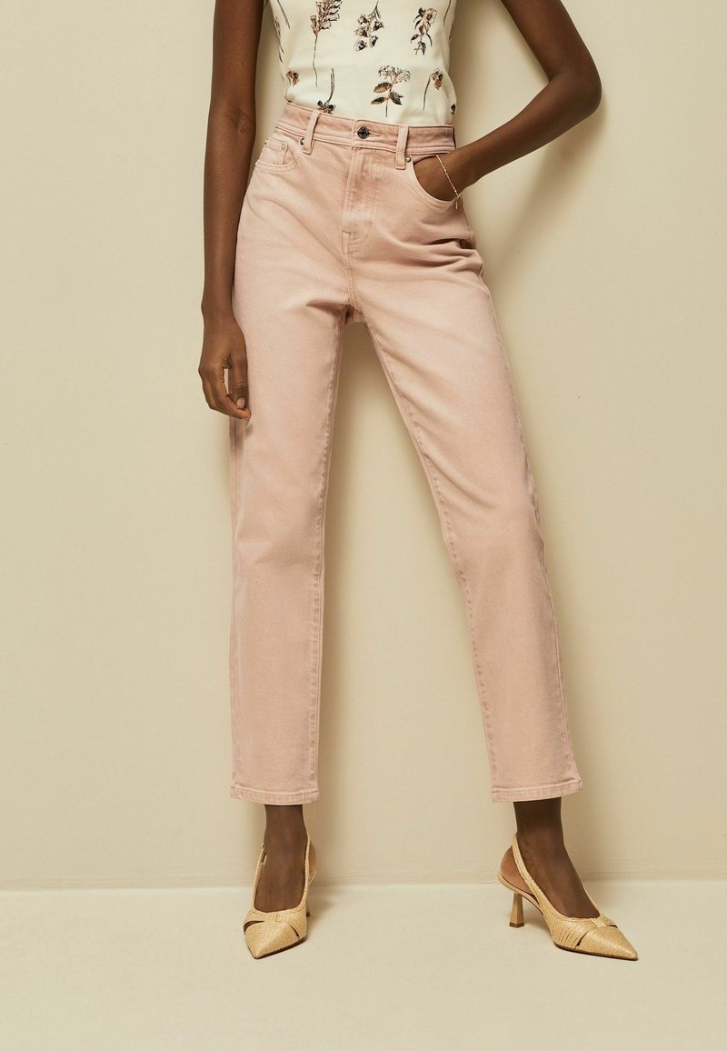 Personne portant un jean taille haute rose clair, un haut blanc sans manches à motif floral et des talons slingback pointus beige, debout contre un mur beige.