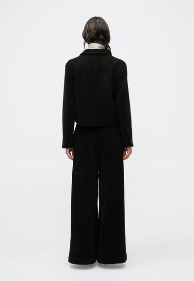 Fekete kordbársony rövidített dzseki bőszárú nadrággal párosítva, puha textúrával és négy zsebbel. Testre szabott kialakítás galléros nyakkivágással.