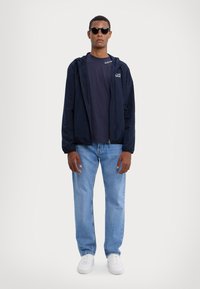 Marineblå vindtæt jakke med hætte, lynlås fortil og EA7-logo; parret med en marineblå T-shirt og lyseblå jeans; hvide sneakers; mørke solbriller.