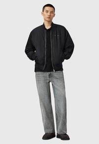 Un jeune homme se tient debout, les mains dans les poches, portant une veste bomber noire, une chemise noire, un jean gris clair et des chaussures foncées sur un fond clair.