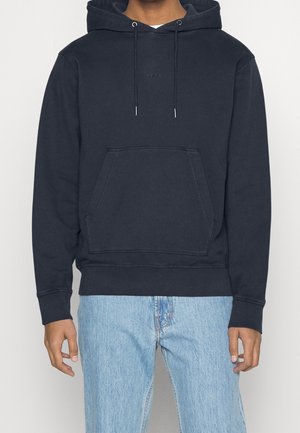 Mann trägt einen schwarzen Kapuzenpullover mit Fronttasche und hellblaue Jeans, steht vor einem hellen Hintergrund.