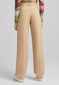 Beige weit geschnittene Hose aus strukturiertem Stoff, mit hohem Bund, Gürtelschlaufen und einer Detailtasche auf der Rückseite.