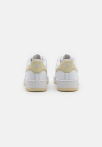Fehér Nike Air tornacipők sima bőrből készült felső résszel, világos sárga részletekkel a saroknál és texturált gumitalppal. A hátsó nézeten a márkajelzés látható.