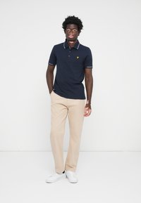 Jūrinio stiliaus polo marškinėliai su pilku apykaklės apdaila ir geltonu logotipu, derinami su beige kelnėmis ir baltais sportbačiais, stovintys prieš neutralų foną.