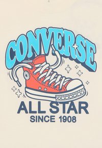 Sarkans Converse kedu zīmējums ar baltām auklām, uz gaismas bēša fona. Teksts: "CONVERSE ALL STAR SINCE 1908."
