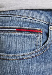 Denim jeans feature a back pocket with red, white, and navy stitching accents, accompanied by a subtle light blue fabric texture.