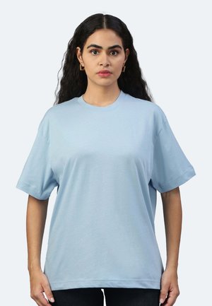Donna con lunghi capelli ricci scuri che indossa una maglietta oversize azzurro chiaro e orecchini quadrati dorati, in piedi contro uno sfondo uniforme.