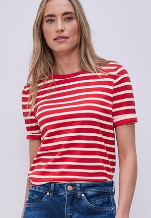 Femme aux longs cheveux blonds portant un t-shirt à manches courtes rayé rouge et blanc, rentré dans un jean en denim bleu, sur un fond uni.