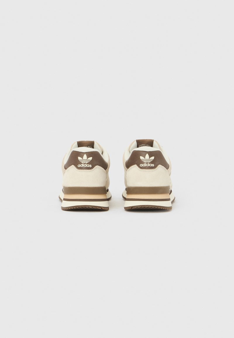 Paire de baskets Adidas beige et marron vues de dos avec logo visible sur la languette marron du talon et semelle rayée sur fond blanc.