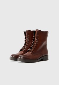 Bottines en cuir marron avec un bout rond, un laçage à l'avant et un petit talon bloc. Texture lisse avec des détails de couture minimaux.