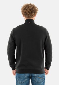 Zwarte sweatshirt met hoge kraag, voorzien van een kleine driekleurige accent bij de hals, gecombineerd met blauwe jeans.