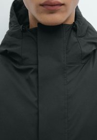 Sort hættejakke med høj krave, glat tekstur, minimalistisk design og syninger langs frontlukningen.