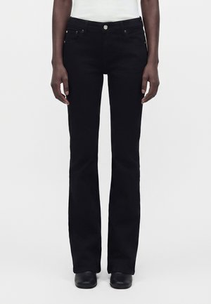 Άτομο που φορά μαύρα bootcut τζιν, λευκό τοπ και μαύρα παπούτσια, στέκεται μπροστά σε μια απλή ανοιχτόχρωμη επιφάνεια.