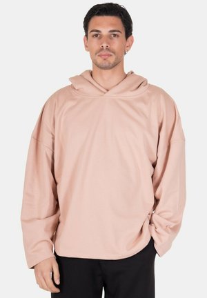Uomo che indossa una felpa beige larga con cappuccio e pantaloni neri, in piedi con le mani rilassate, espressione neutra su sfondo bianco.