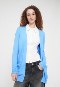 Ανοιχτό γαλάζιο ζακέτα από μαλακό πλεκτό ύφασμα, με ανοιχτό μπροστινό μέρος, ριμπ μανσέτες και δύο πλευρικές τσέπες, φορεμένη πάνω από ένα λευκό πουκάμισο με κουμπιά.