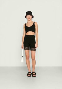 Donna che indossa un crop top senza maniche nero, pantaloni corti neri a vita alta con bordo in pizzo, cappello a secchiello nero, sandali neri e tiene in mano una piccola borsa bianca.