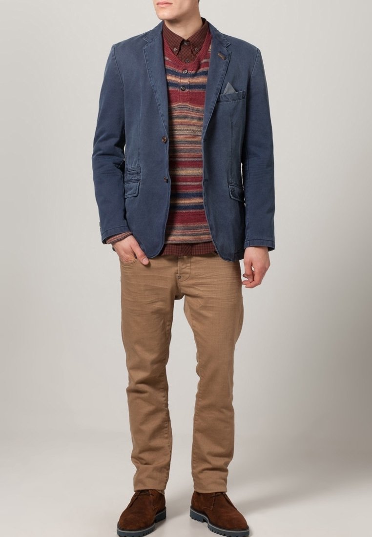 Homme portant un blazer bleu, un pull rayé multicolore, une chemise marron, un pantalon beige et des chaussures marron, debout avec une main dans la poche.