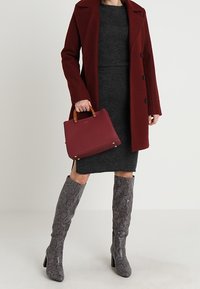 Cappotto bordeaux, vestito grigio, stivali grigi a ginocchio con motivo e borsa bordeaux strutturata con manici in legno.