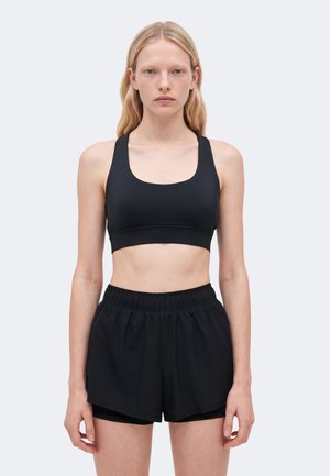 Una mujer luce un sujetador deportivo negro y unos pantalones cortos deportivos negros y holgados sobre un fondo claro y liso.