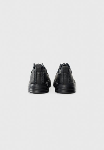 Pair of black textured leather sneakers with thick ridged soles, viewed from the back on a plain light grey background.