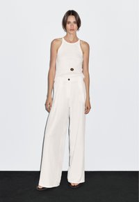 Witte mouwloze top en wijde broek, gemaakt van lichtgewicht stof. Bevat een zwarte knop en subtiele zakken. Eenvoudige lange ketting.