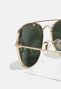 Ochelari de soare aviator cu ramă de aur și lentile de un verde închis. Caracteristici includ brațe metalice subțiri și suporturi pentru nas transparente. Formă clasică și finisaj lucios.