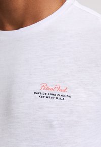 T-shirt bianco a maniche corte realizzato in morbido cotone, con logo rosa "Petrol Ind." e testo nero "Bayside Lane Florida Key-West U.S.A."